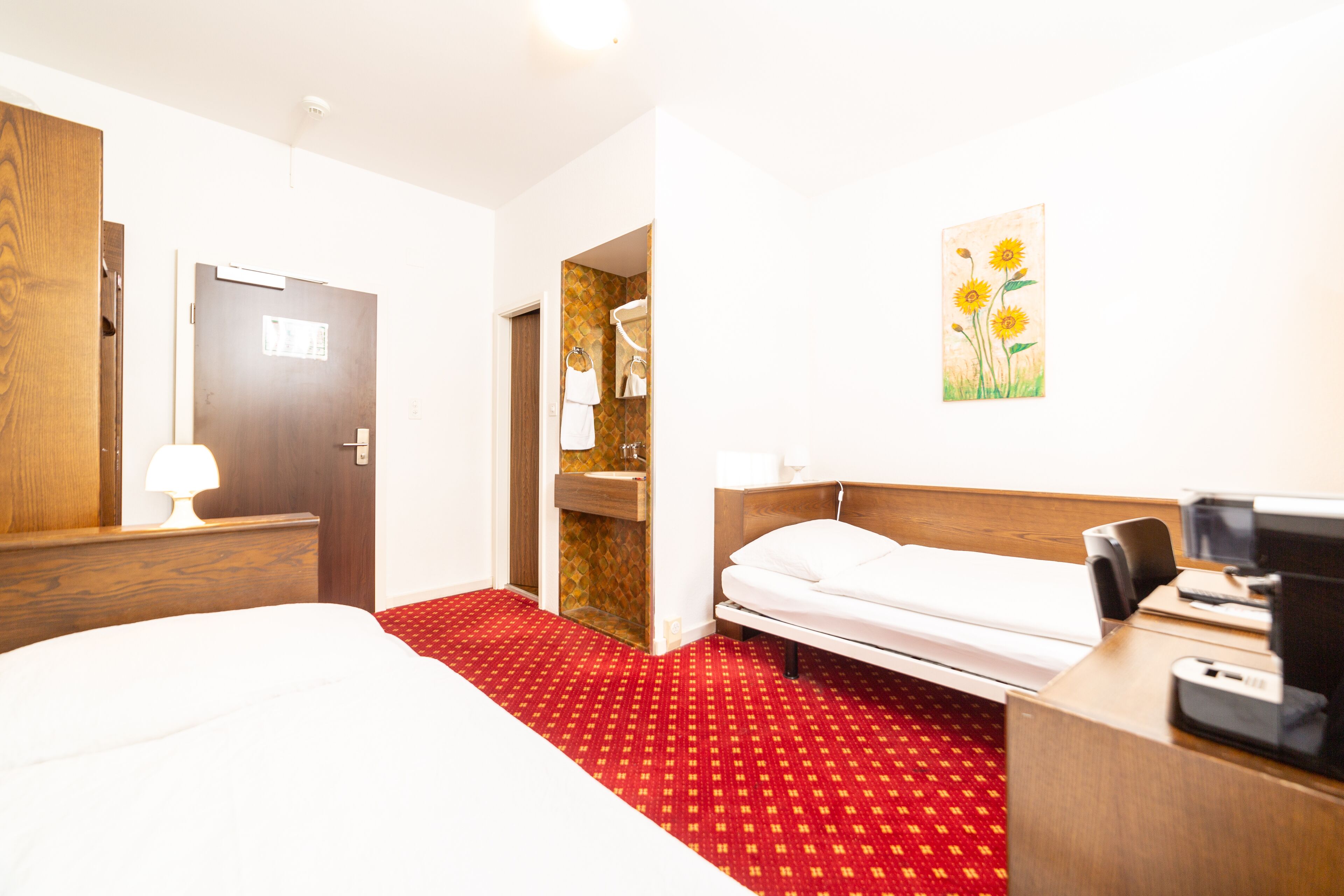 A hotel room with a bed, a desk with a computer, a mirror, and a red carpet.