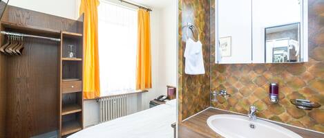 A hotel room with a bed, a wooden wardrobe, a bathroom with a sink and mirror, and a window with orange curtains.