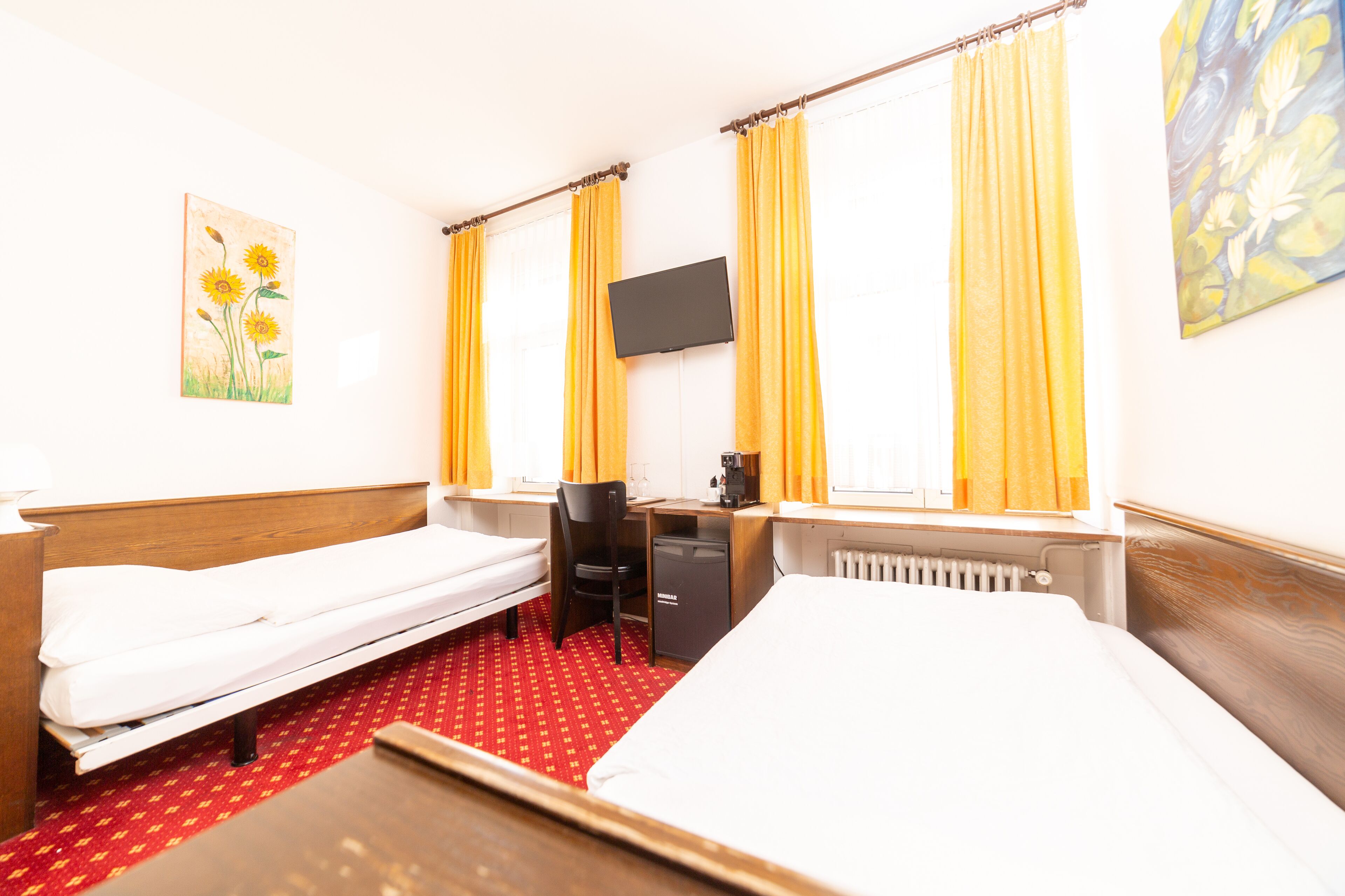 A hotel room with two single beds, a desk with a chair, a TV mounted on the wall, and a painting of flowers.