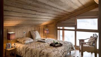 Een gezellige slaapkamer met een houten plafond, een groot bed met bloemenbeddengoed, een nachtkastje met een lamp en een stoel bij een raam.