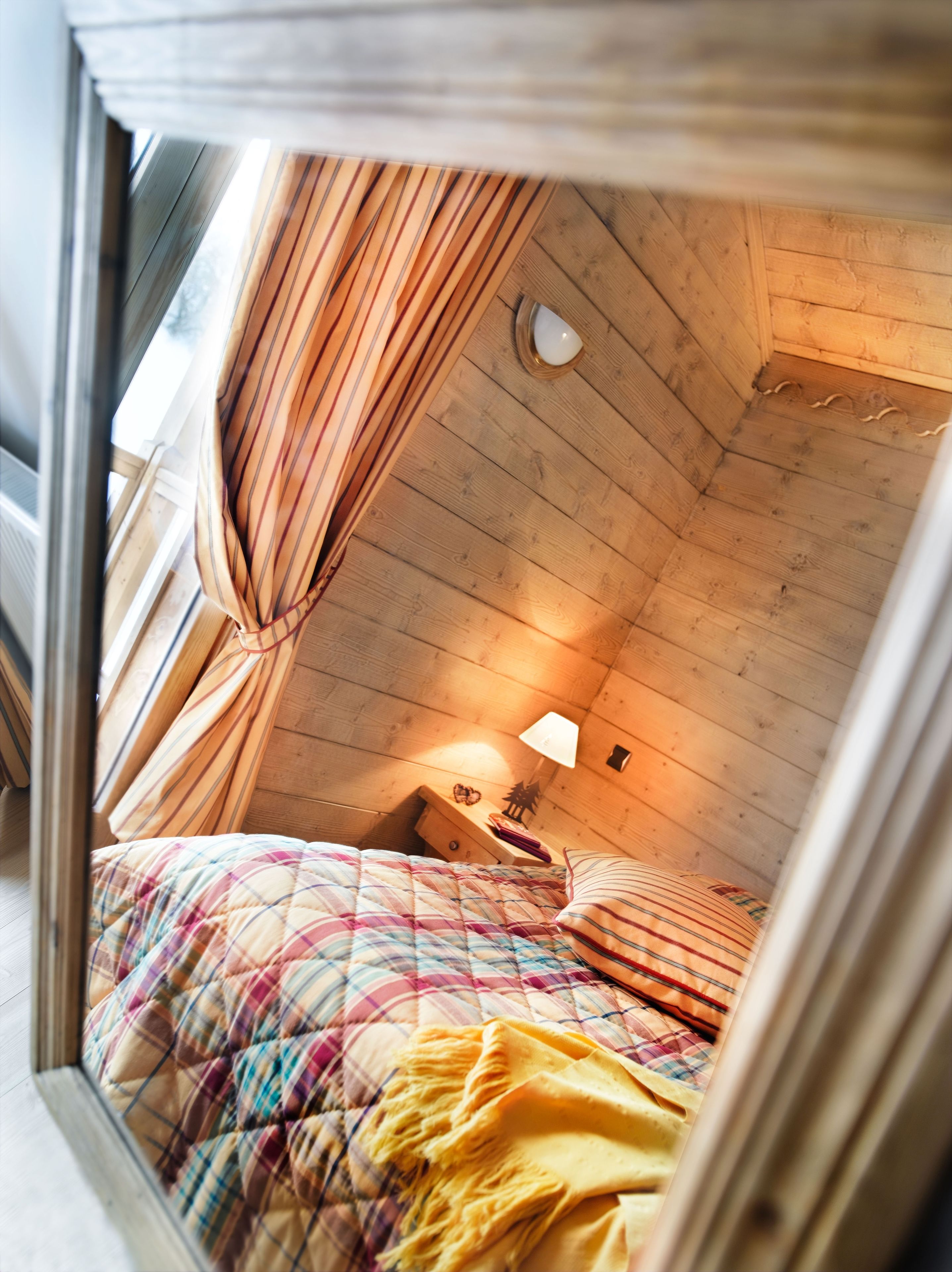A cozy attic bedroom with a wooden sloped ceiling, a bed with a plaid comforter, a bedside lamp, and a window with curtains.