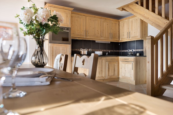A wooden kitchen with cabinets, a stove, and a dining table set with glassware. - CGH Résidences & Spas Village de Lessy (Le Grand-Bornand)