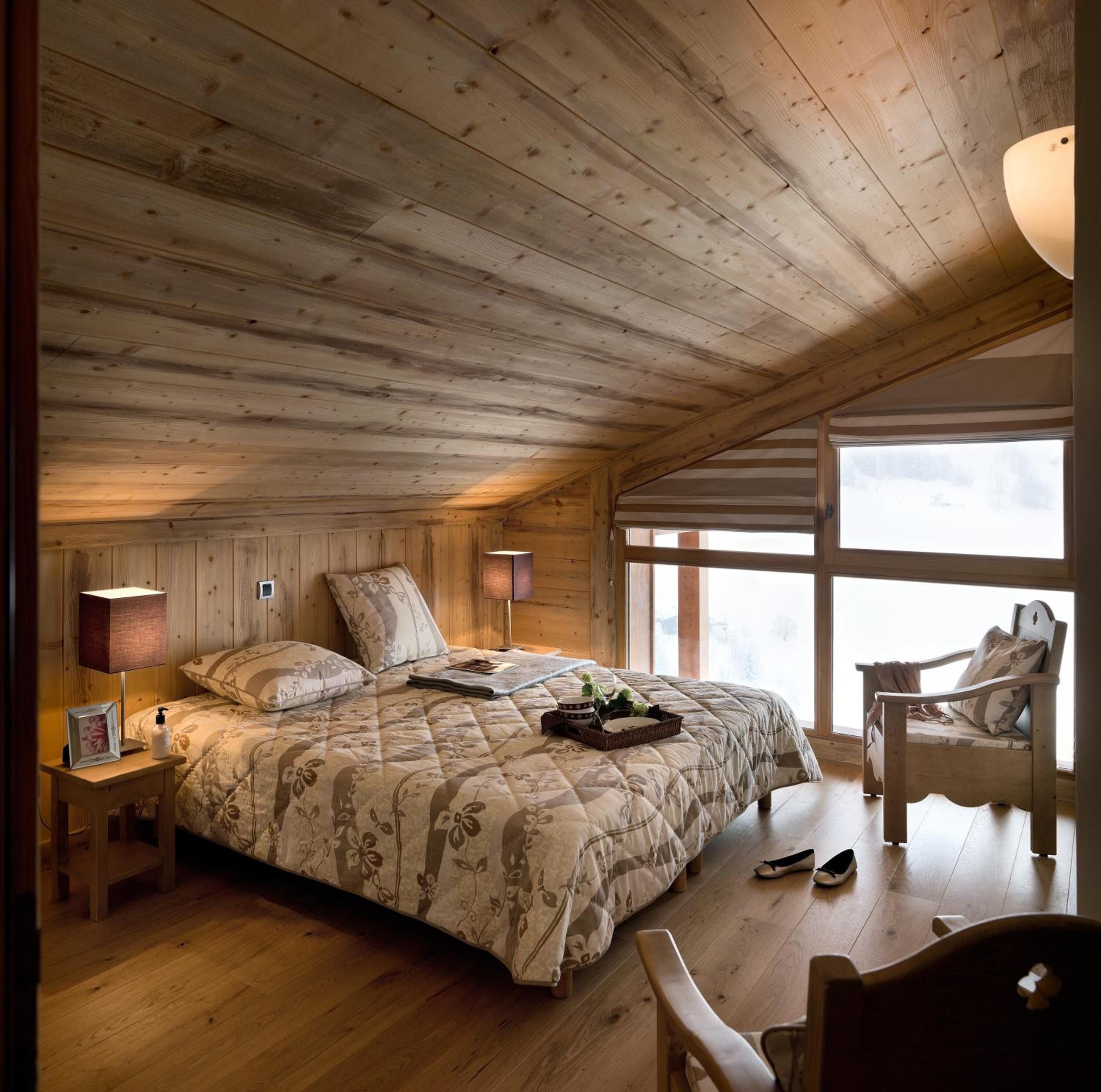 Ein gemütliches Schlafzimmer mit Holzbalkendecke, einem großen Bett mit floralem Bettzeug, Nachttischlampen, einem Sessel und Blick ins Freie.