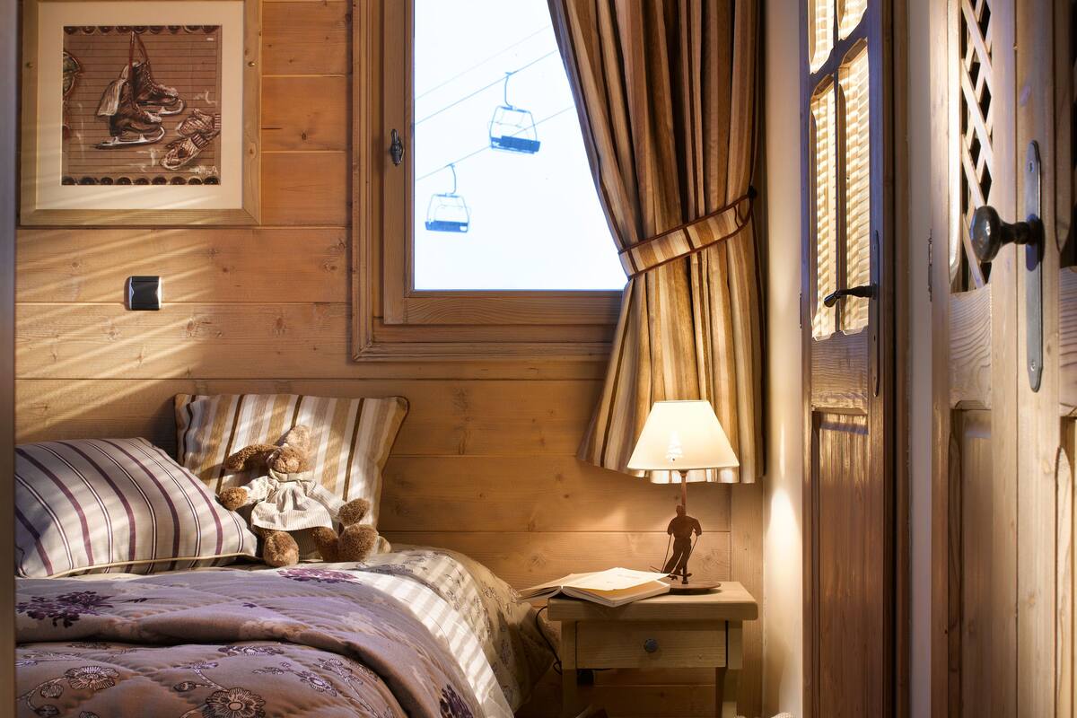 Un dormitorio acogedor con techo de madera, una ventana con vista a un telesilla, una cama con ropa de cama floral, una mesita de noche con una lámpara y un osito de peluche.