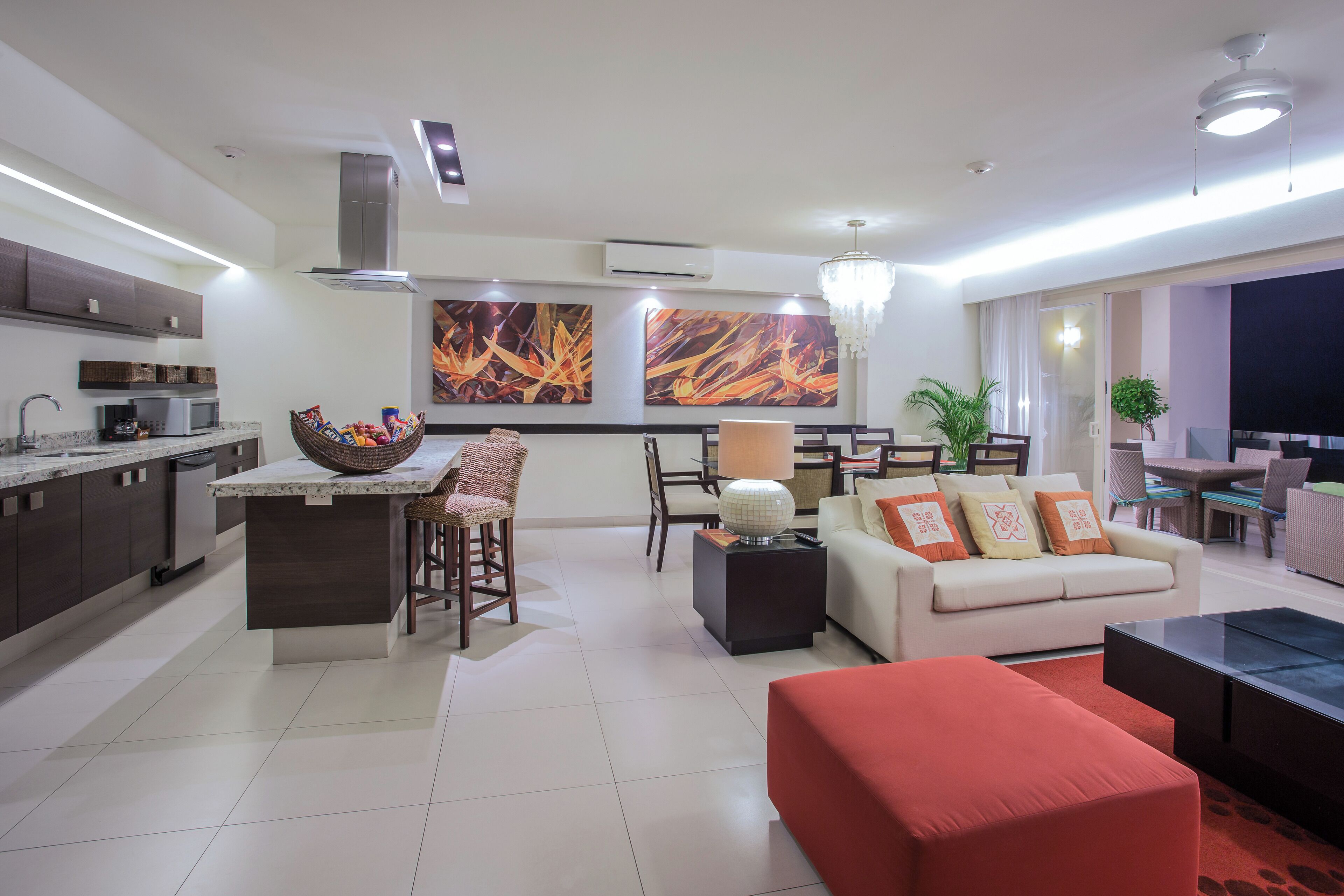 Una sala moderna con sofá, un reposapiés rojo, una zona de comedor con mesa y sillas, y una cocina con microondas y un cesto de aperitivos.