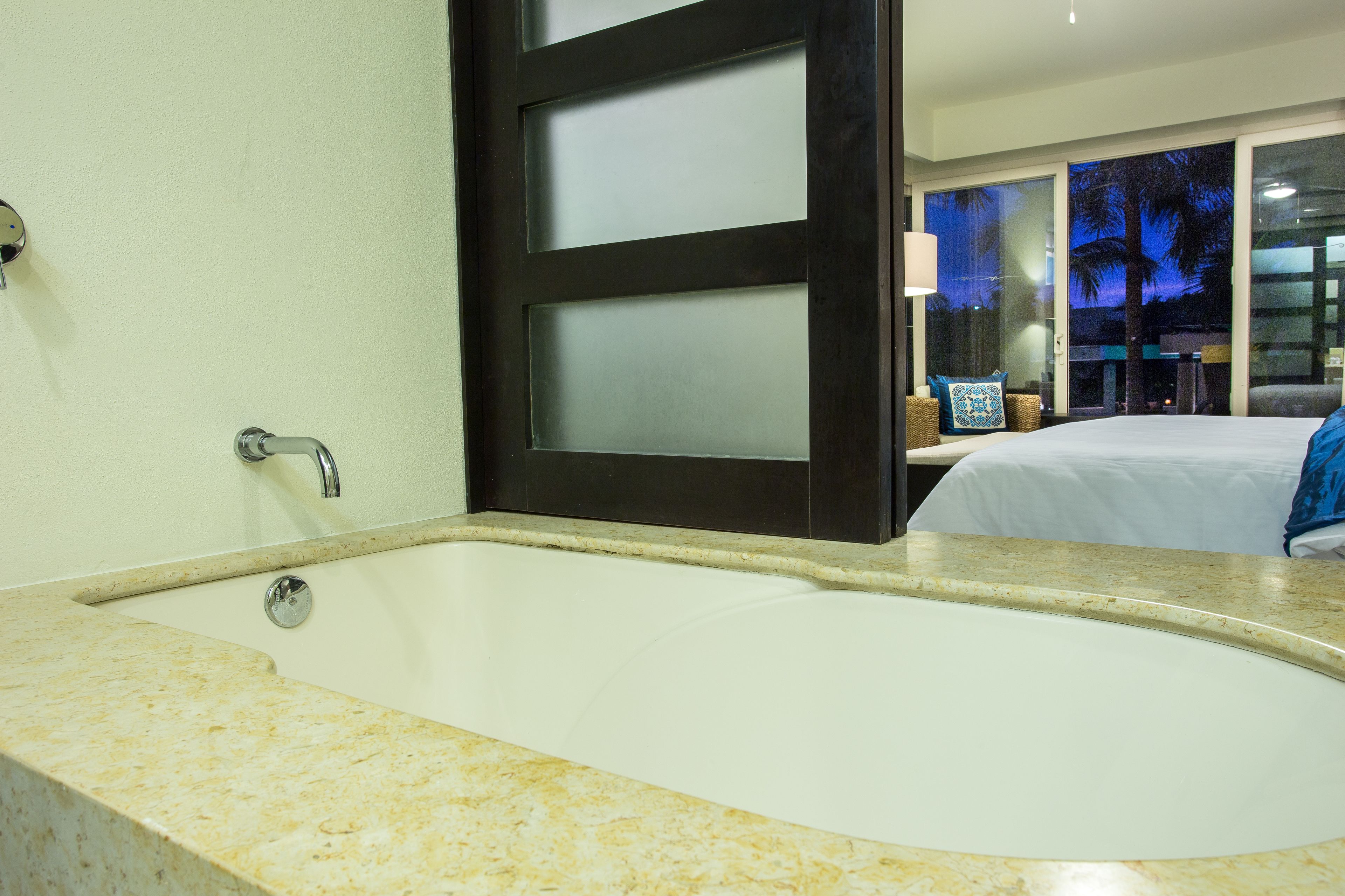 A bathroom with a large bathtub, a door with frosted glass panels, and a view of a balcony with a bed and a lamp.