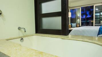A bathroom with a large bathtub, a door with frosted glass panels, and a view of a balcony with a bed and a lamp.