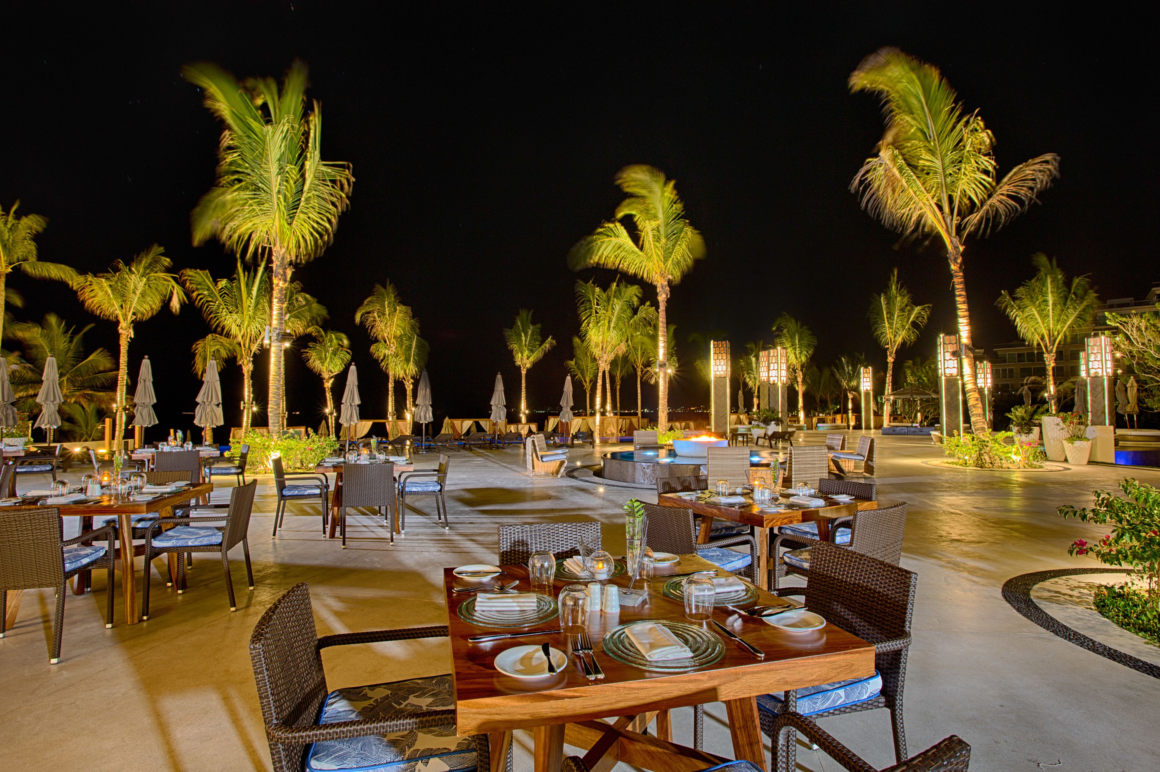 An outdoor dining area with tables and chairs, palm trees, and ambient lighting.