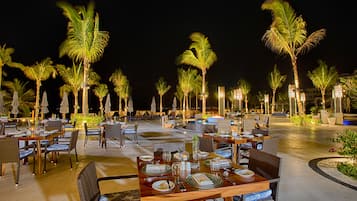 An outdoor dining area with tables and chairs, palm trees, and ambient lighting.