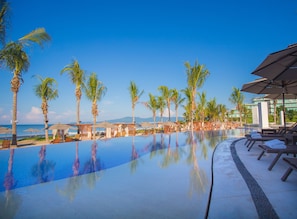 Zona junto a la piscina con sillones de descanso y sombrillas, palmeras y vista al océano.