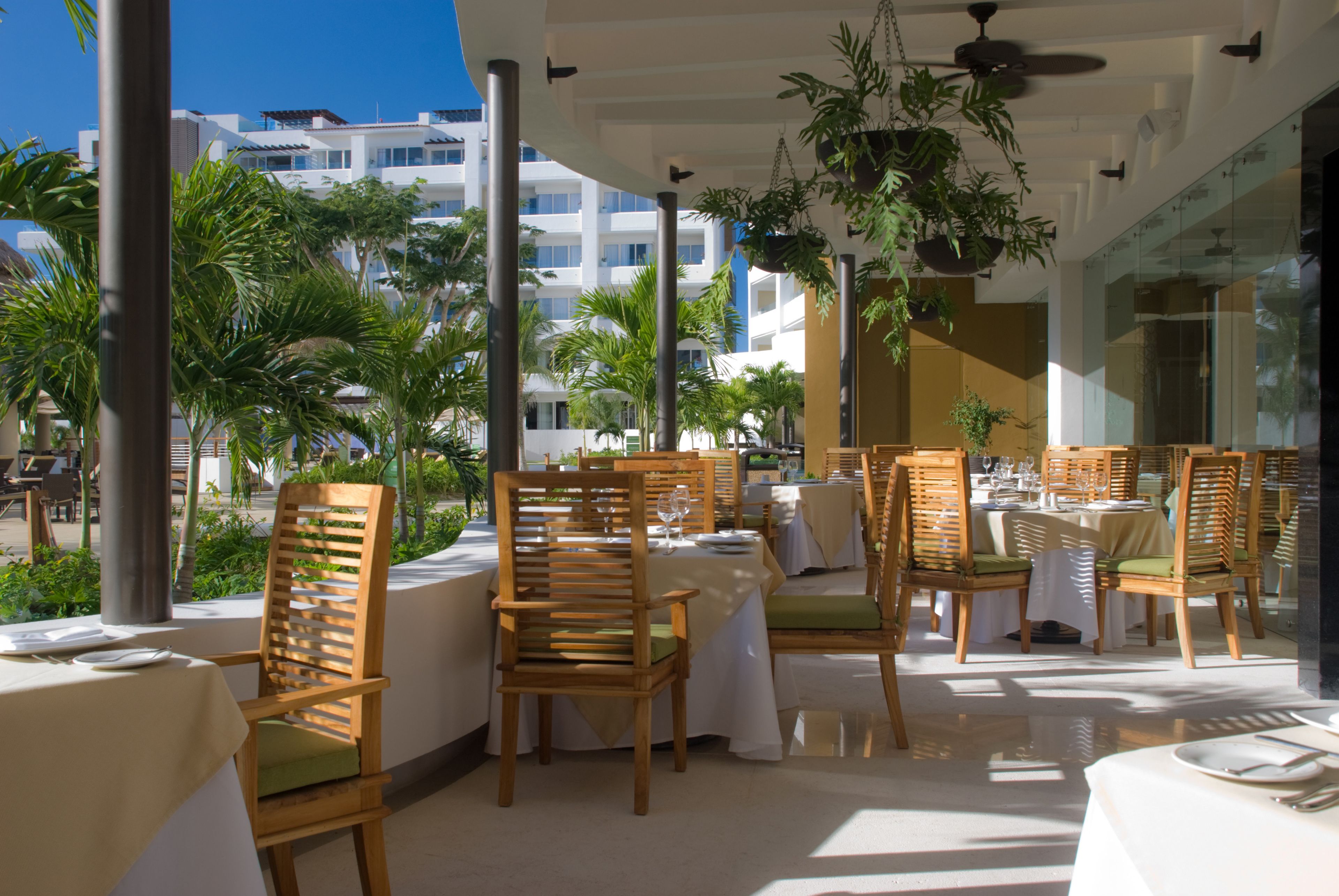 An outdoor dining area with wooden chairs and white tablecloths, overlooking a pool and palm trees.