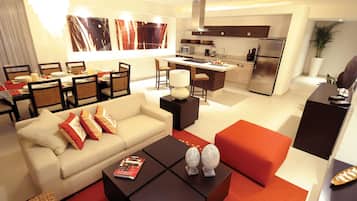 Una sala de estar moderna con un sofá, una mesa de centro y un otoman rojo. Se observa un comedor con una mesa y sillas. El área de la cocina incluye un refrigerador, microondas y un lavamanos.