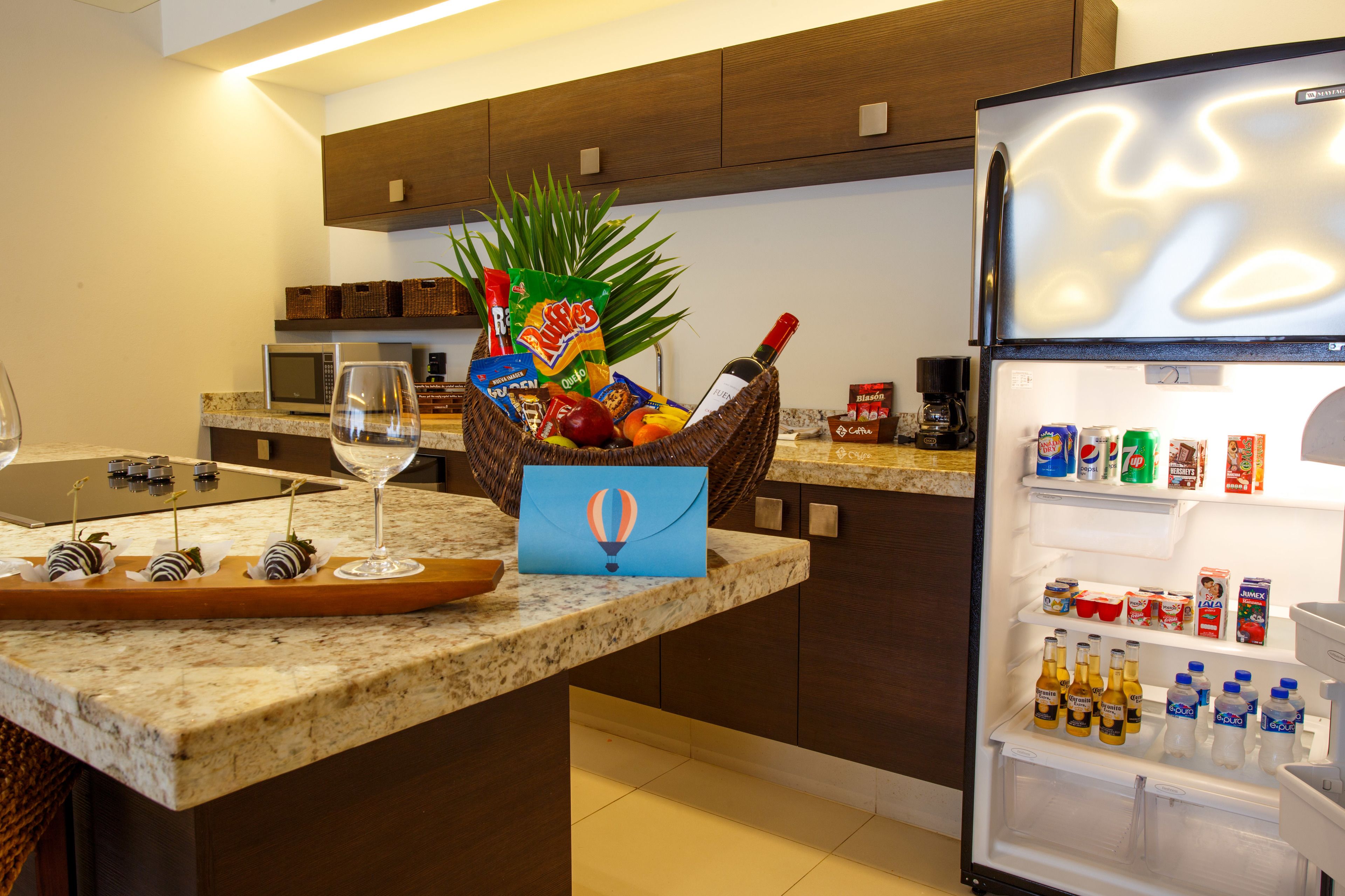 A kitchen with a marble countertop, a wicker basket with snacks, a wine glass, and a refrigerator stocked with beverages.