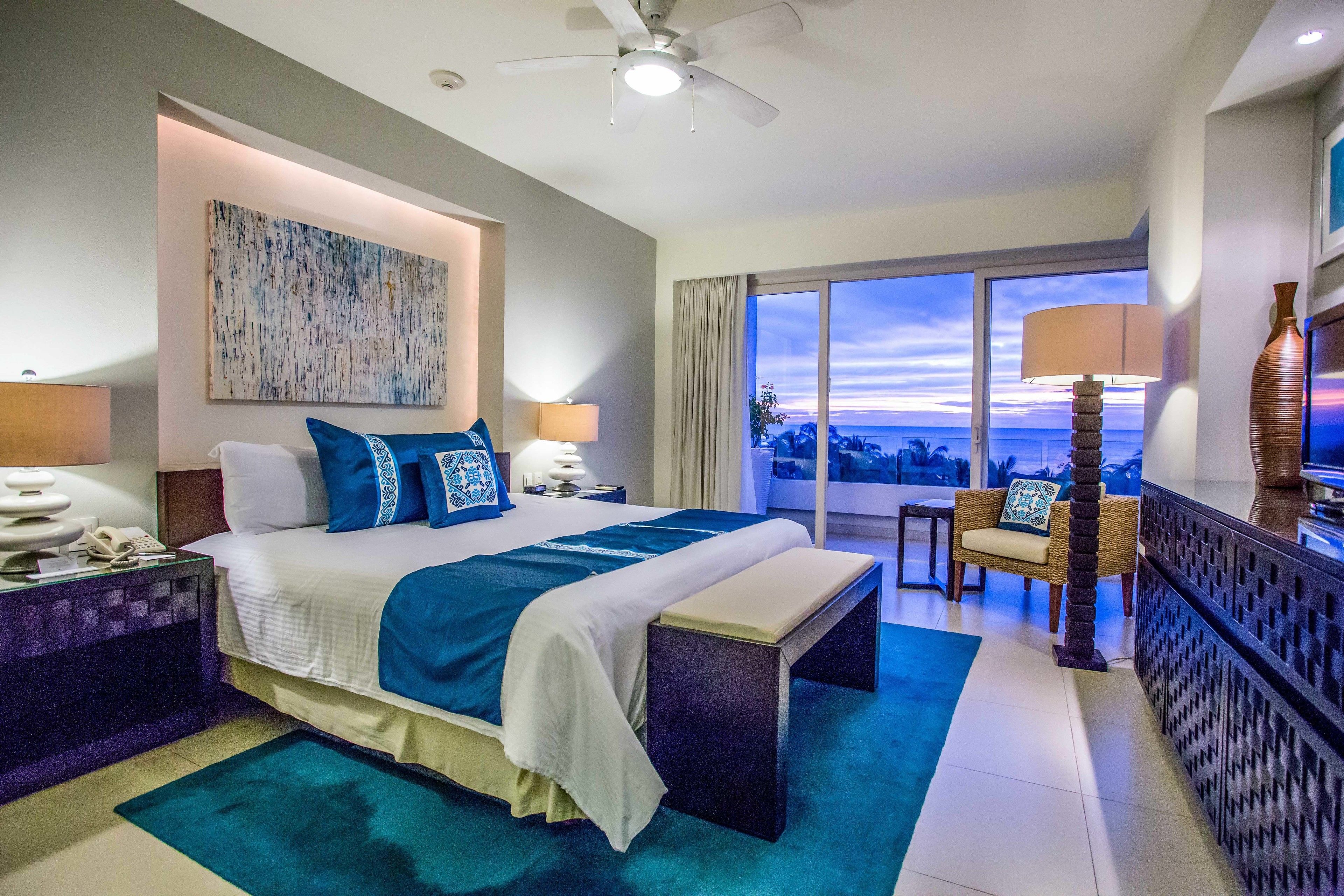 Quarto de hotel com uma cama grande, uma escrivaninha, uma cadeira, varanda com vista e um ventilador de teto.