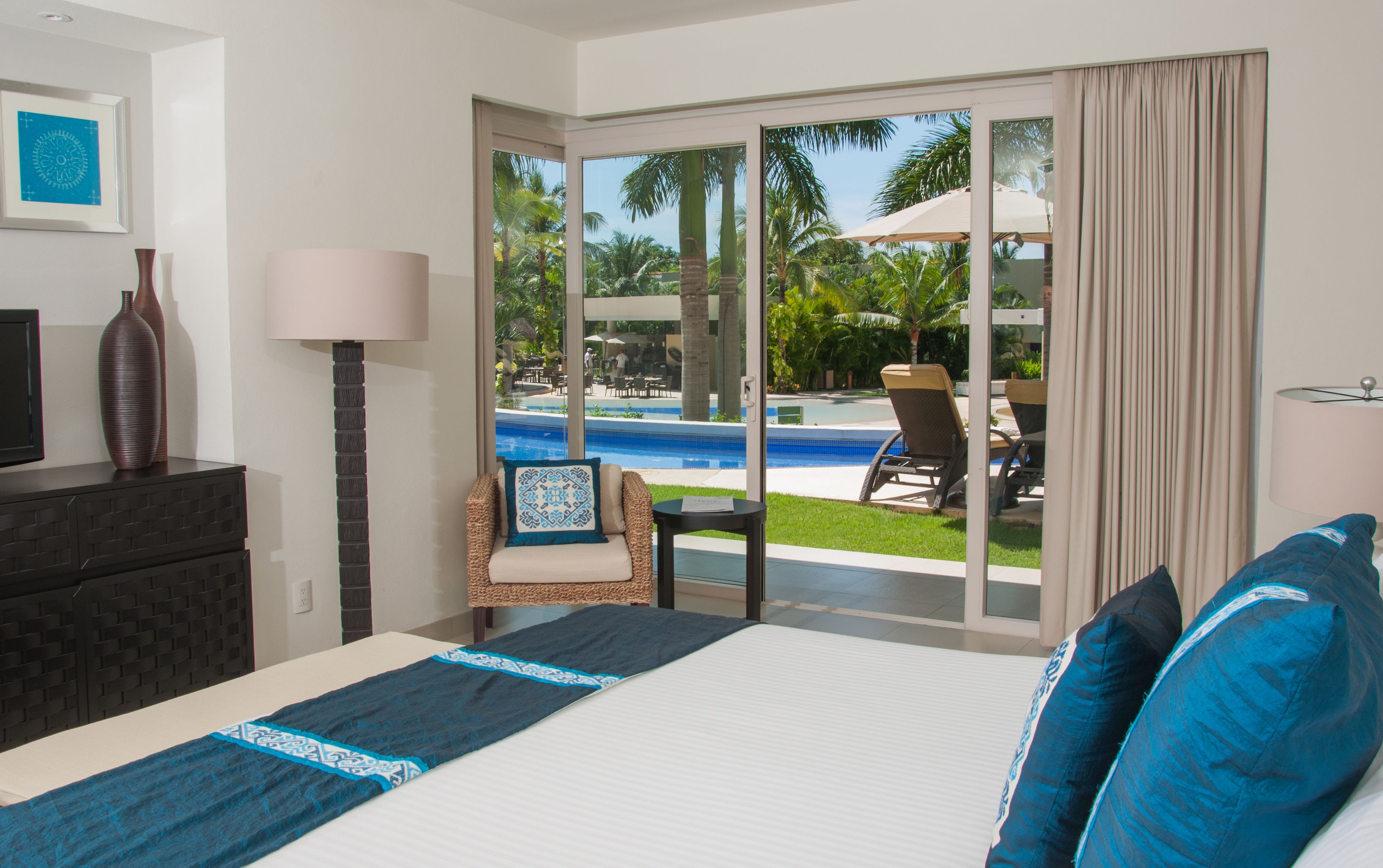 A hotel room with a bed, a chair, a desk, a TV, and a view of a pool and palm trees.