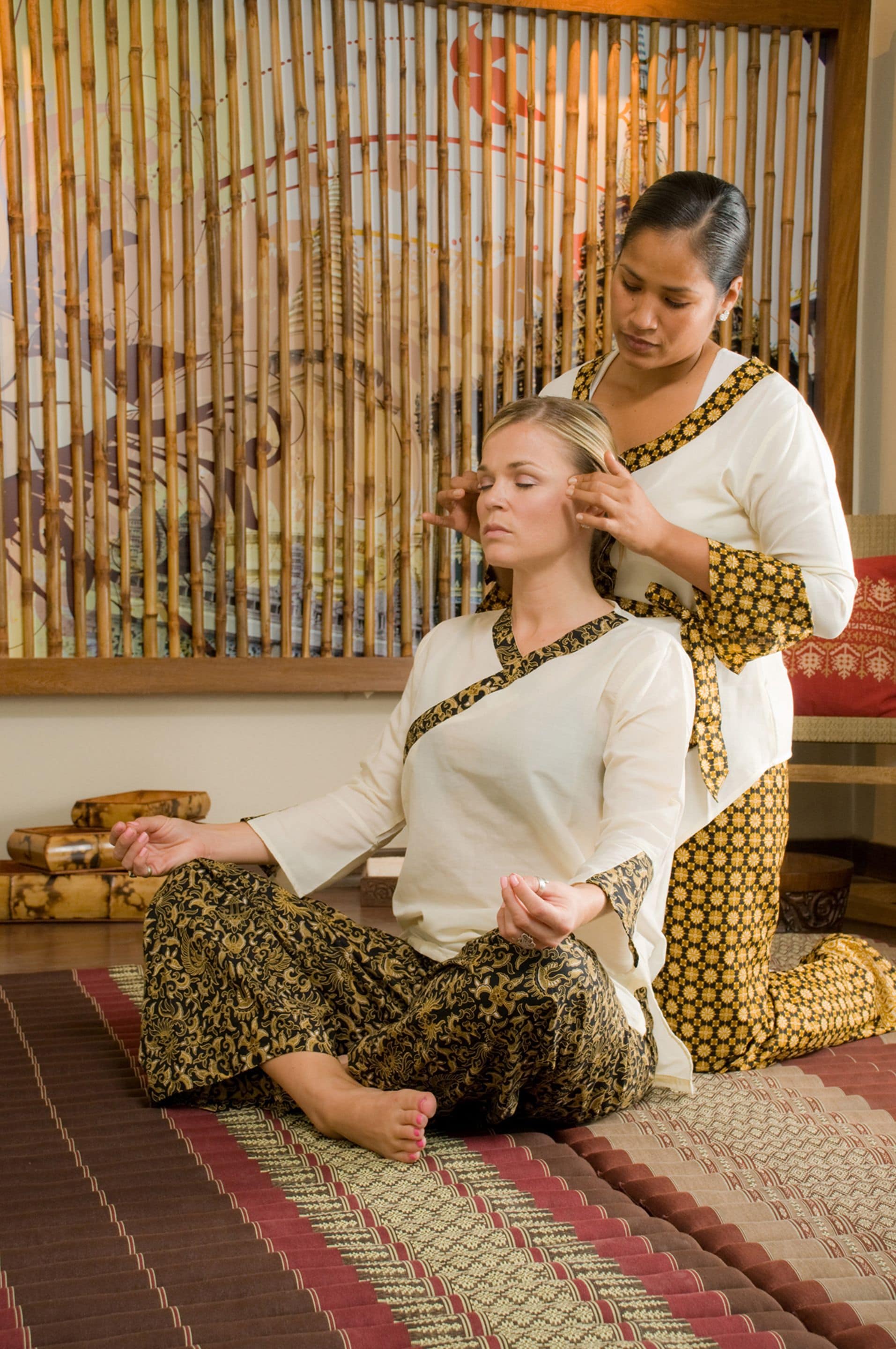 A person sitting on a patterned mat in a room with traditional decor, including a bamboo screen and a red cushion.