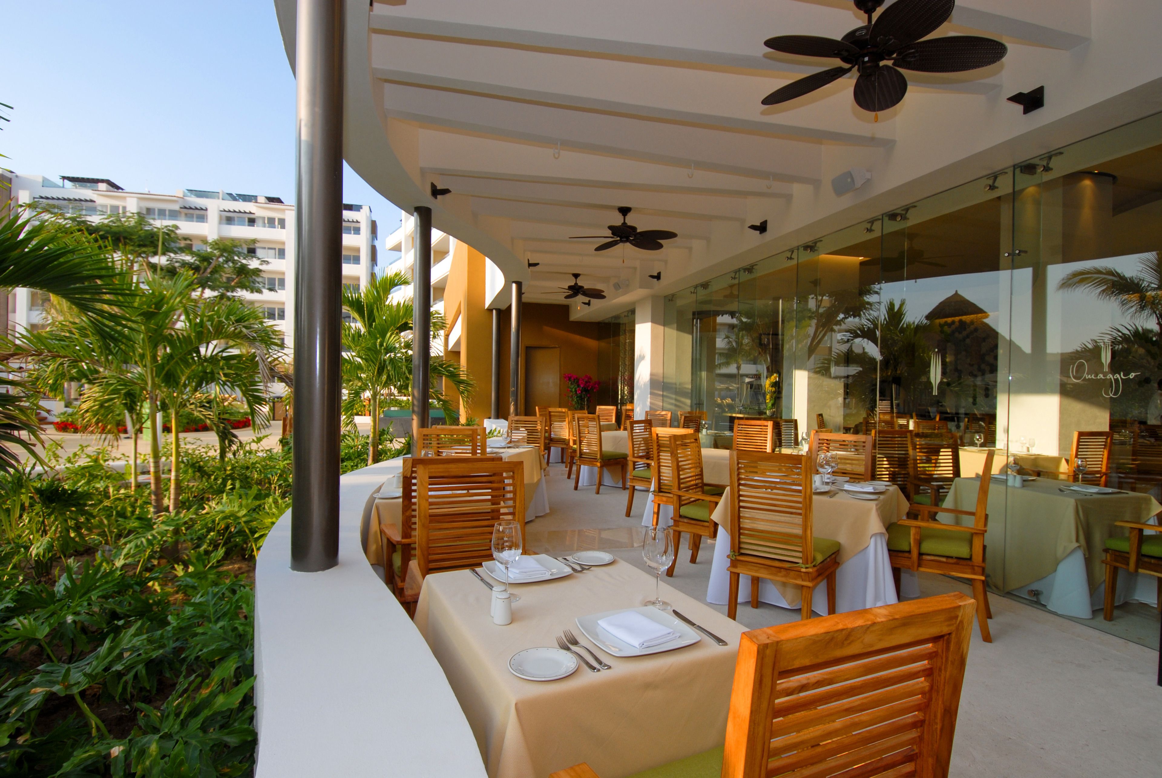 An outdoor dining area with tables and chairs, ceiling fans, and a view of palm trees and buildings.