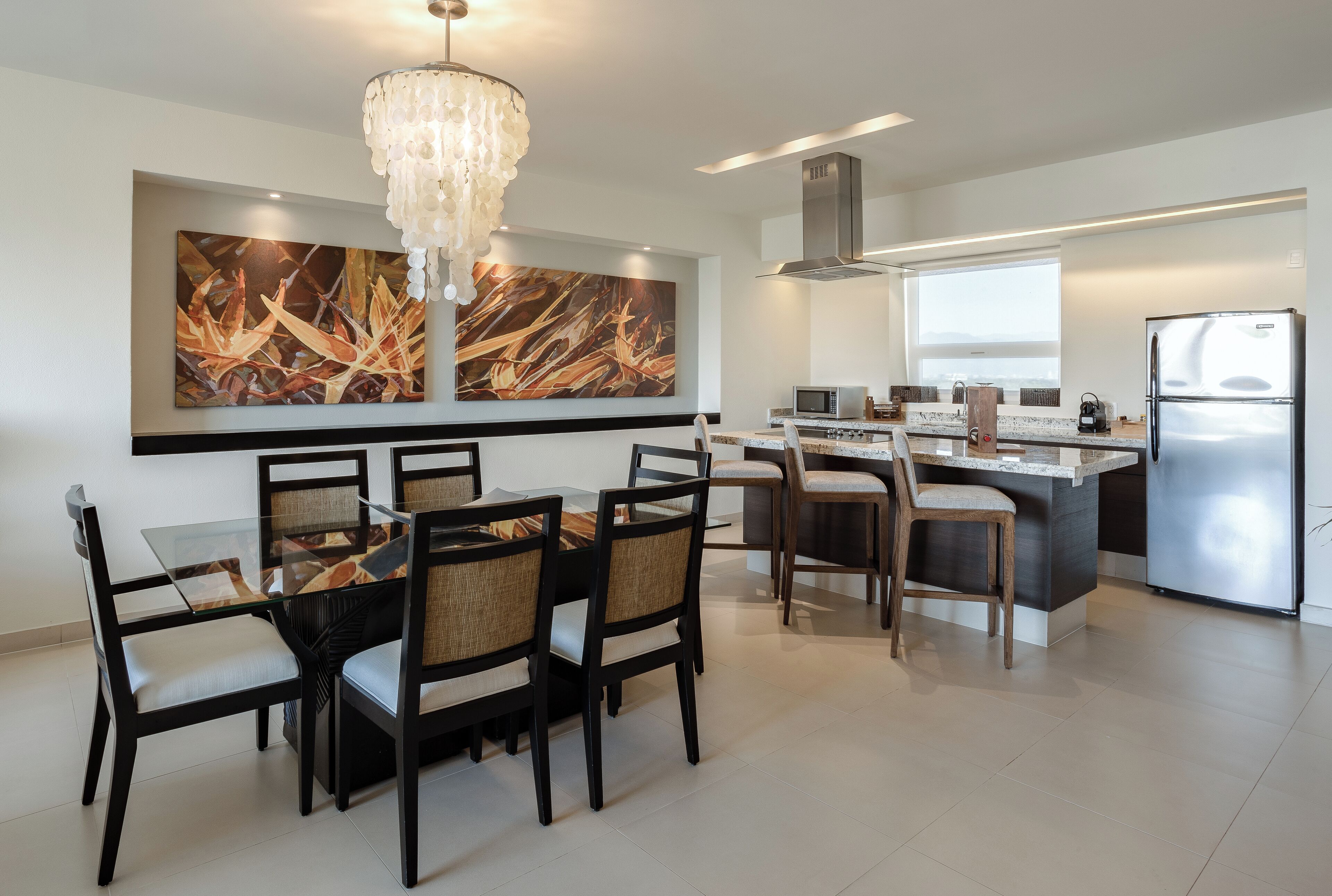 A modern kitchen with a dining area, featuring a chandelier, a large abstract painting, and a stainless steel refrigerator.