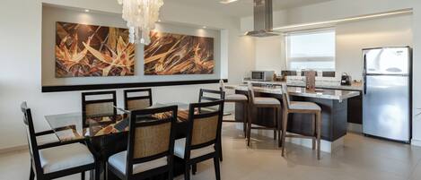A modern kitchen with a dining area, featuring a chandelier, a large abstract painting, and a stainless steel refrigerator.