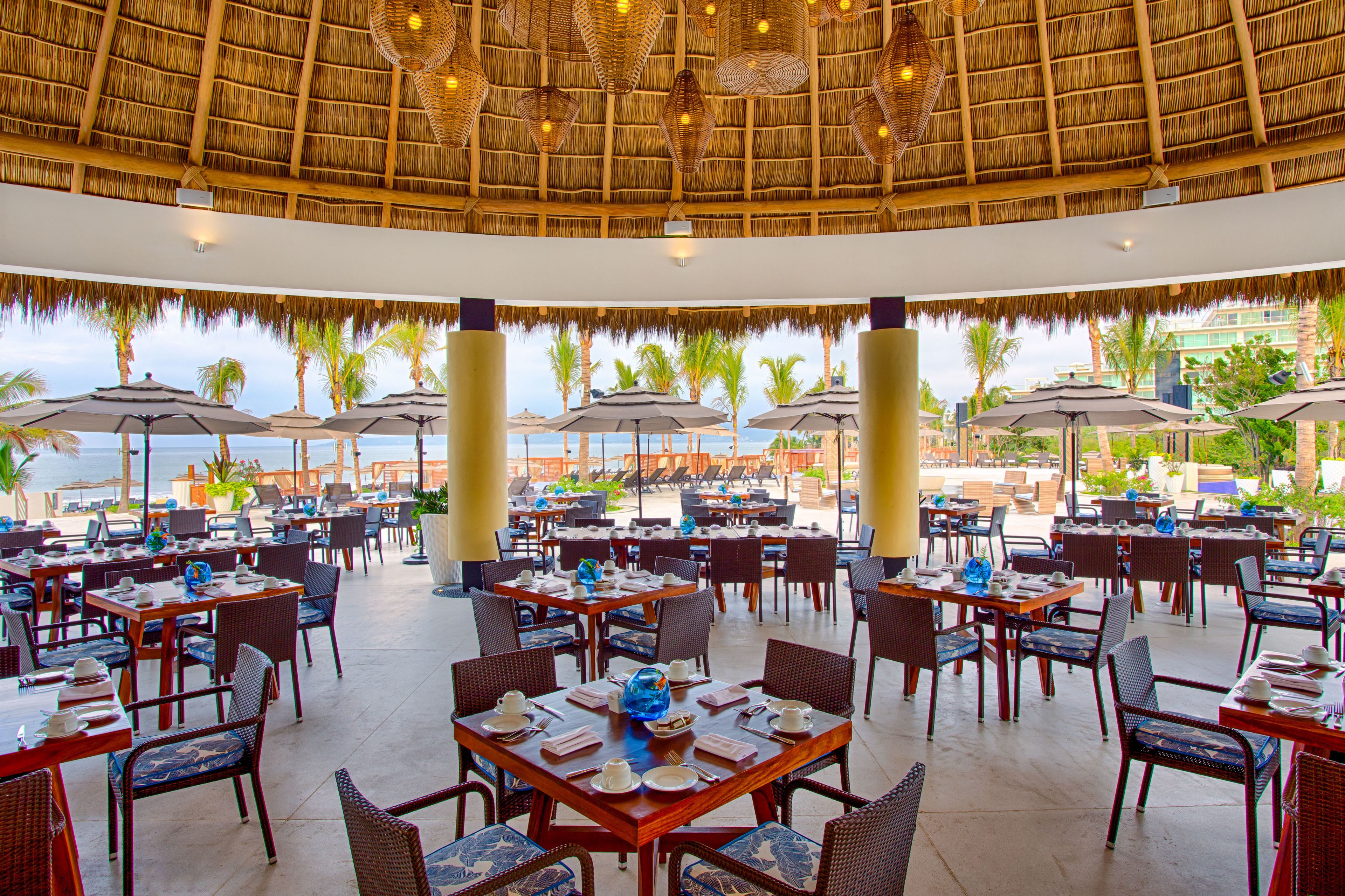A restaurant with tables and chairs, a thatched roof, and a view of the ocean.