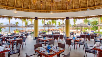 A restaurant with tables and chairs, a thatched roof, and a view of the ocean.