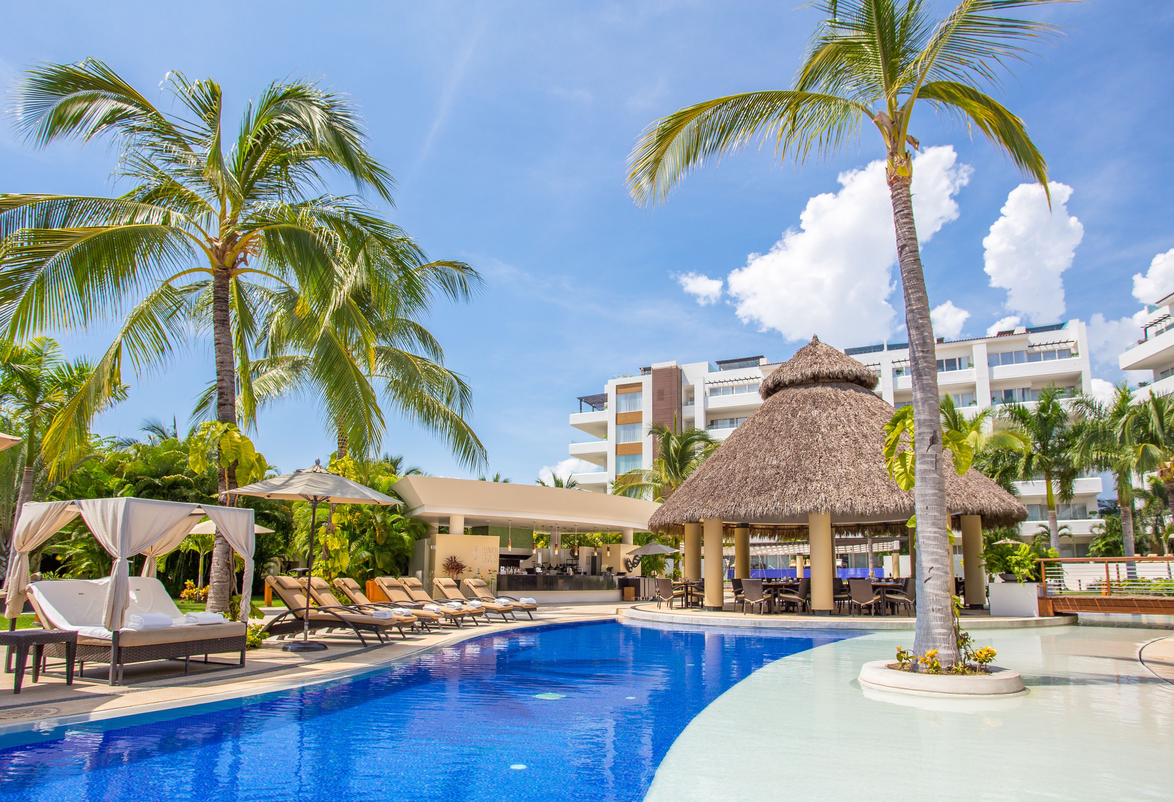 A resort pool area with lounge chairs, a thatched-roof gazebo, and palm trees.