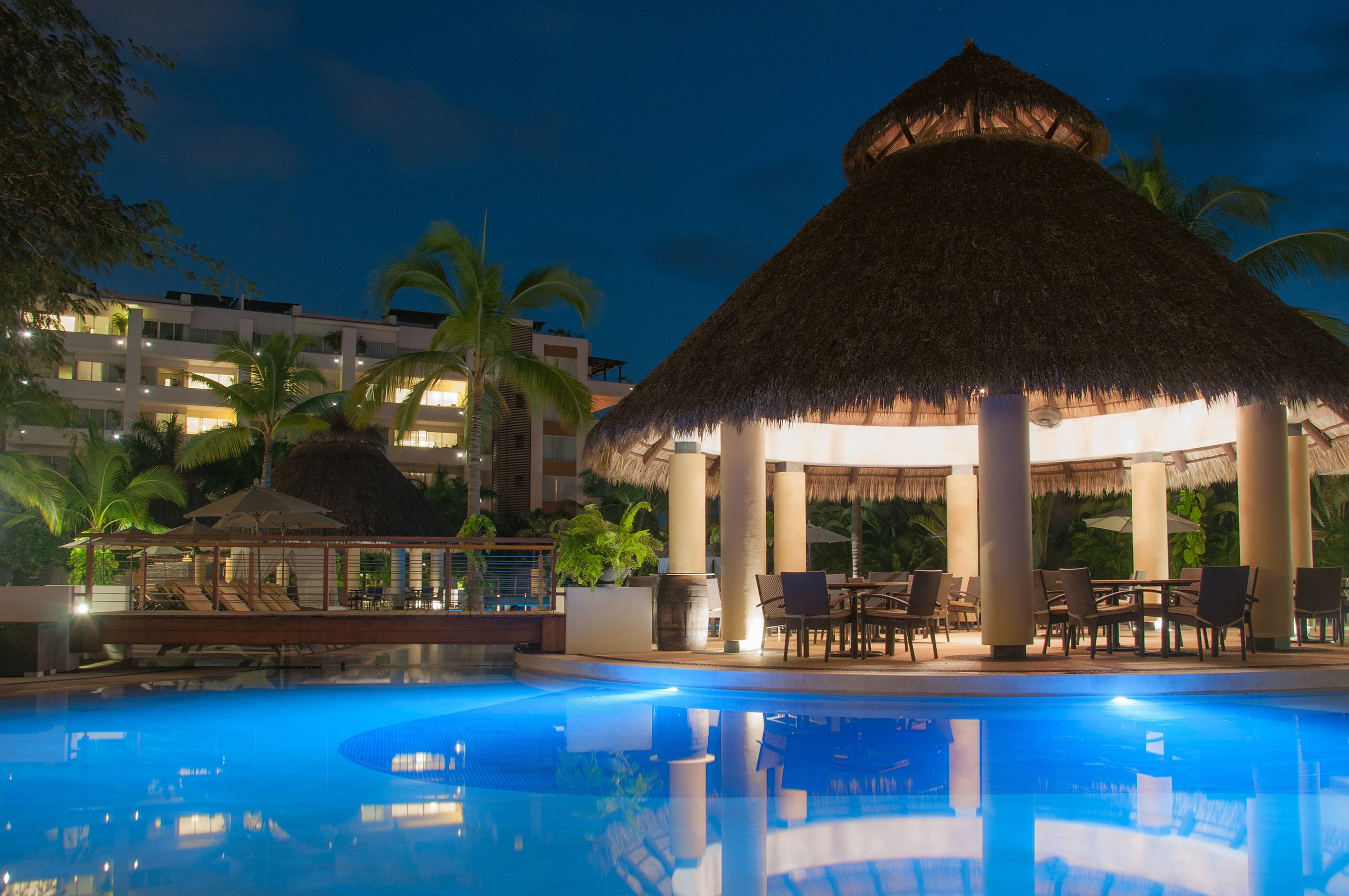 A swimming pool with a thatched-roof pavilion, lounge chairs, and palm trees.