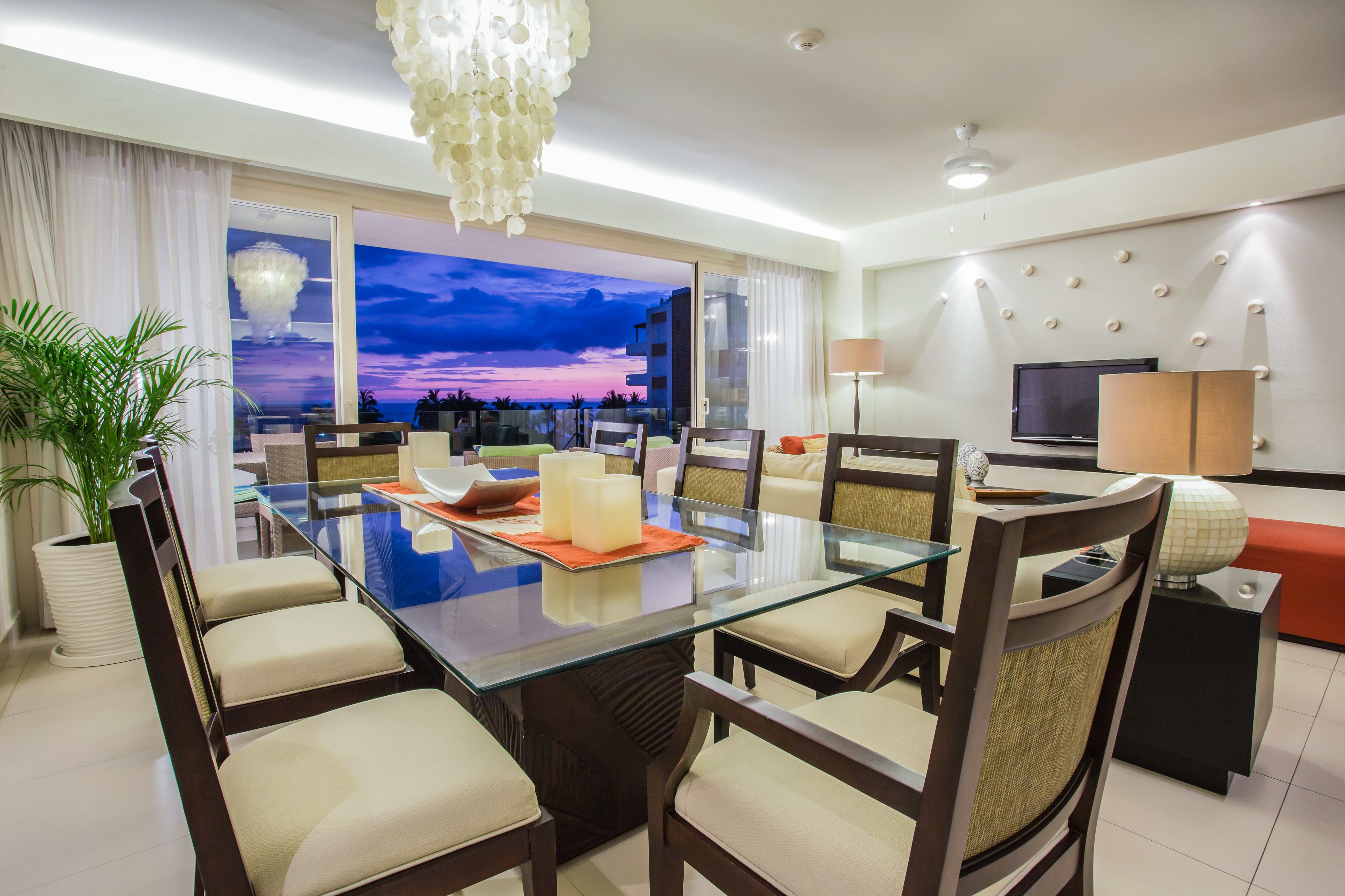 A modern hotel room with a glass dining table, comfortable seating, a large window with a sunset view, a flat-screen TV, and a decorative wall.