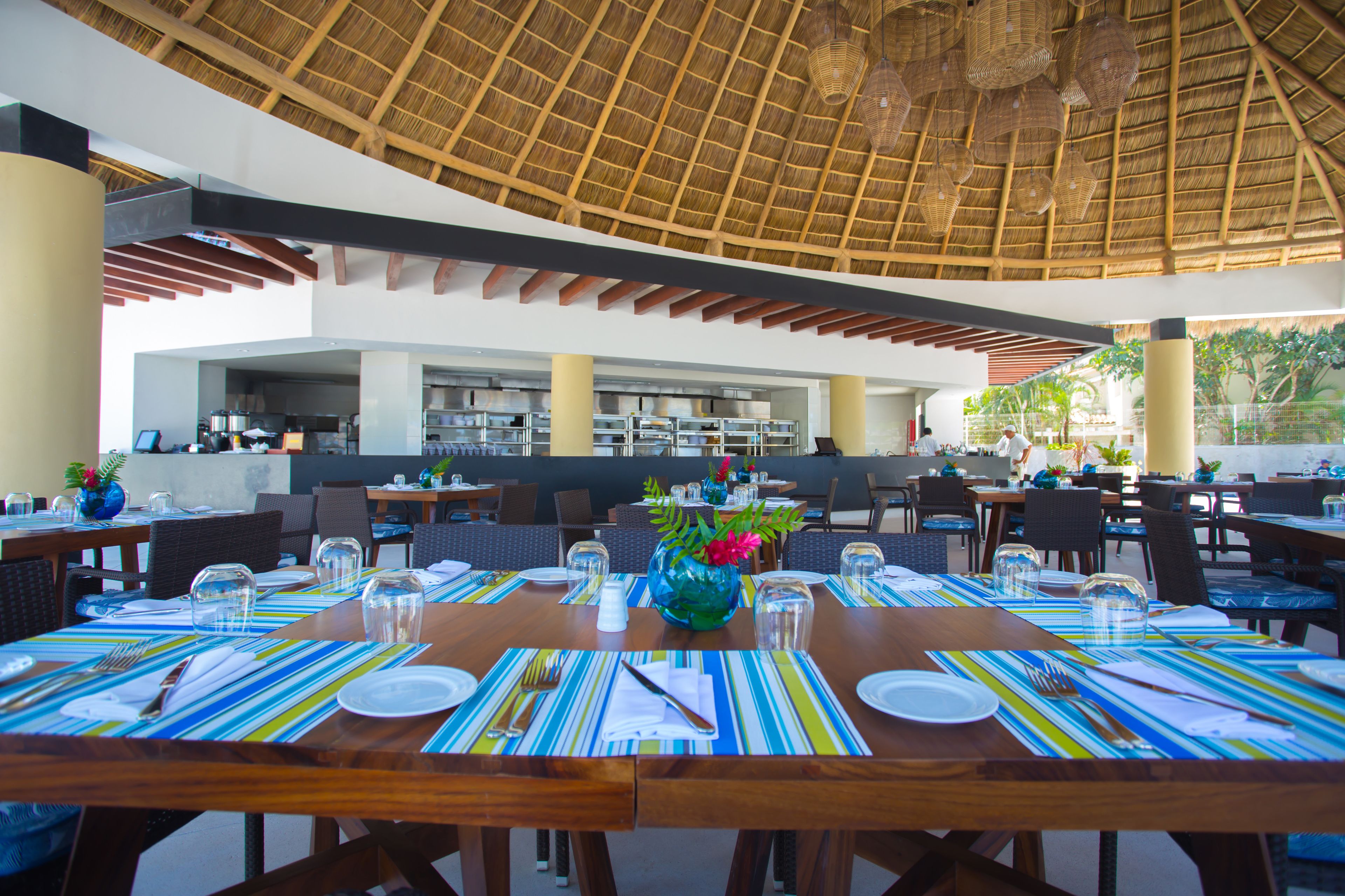 Un restaurant avec des tables et des chaises en bois, un toit de chaume et une vue sur l’extérieur.