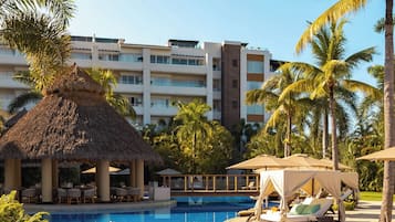 Zona de piscina en un resort con una estructura de techo de paja, sillas de descanso y palmeras.