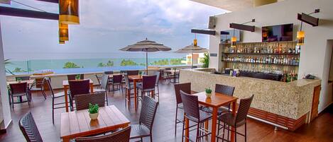 A rooftop bar with wooden tables and chairs, umbrellas, and a bar counter with a view of the ocean.