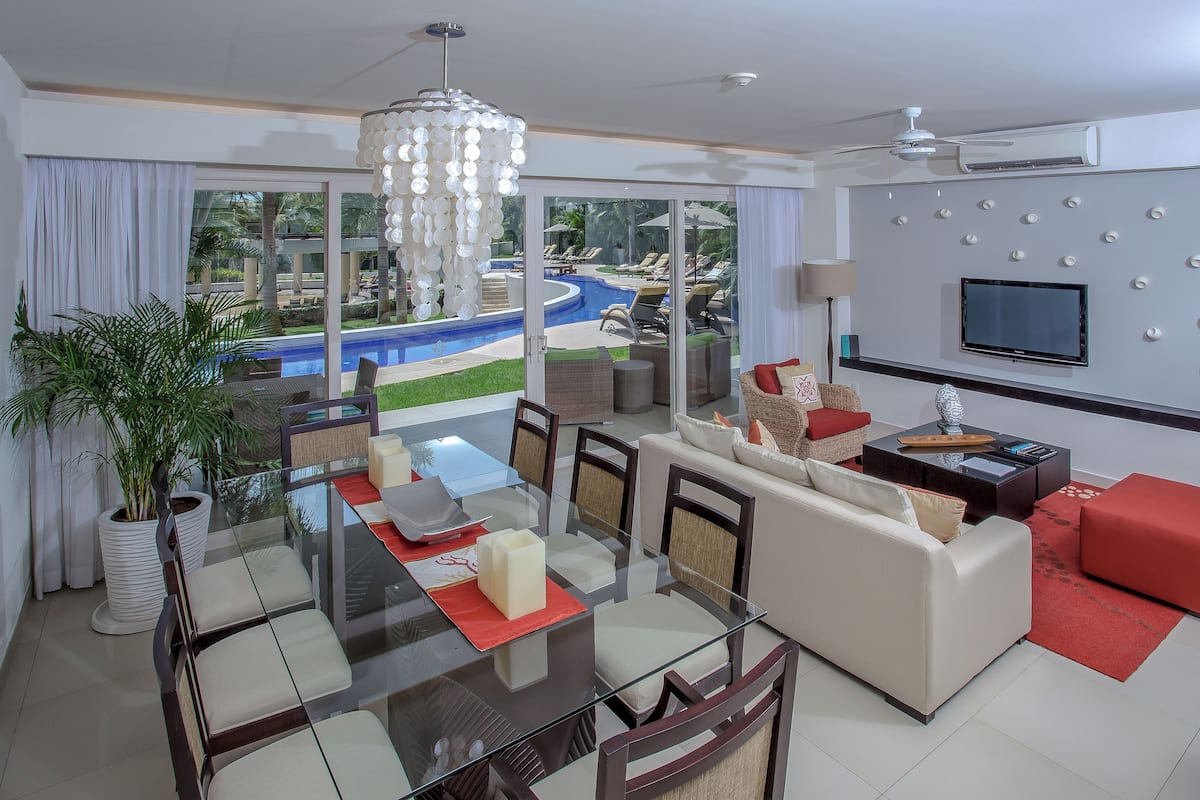 Ein modernes Wohnzimmer mit einem Glastisch, bequemen Sitzgelegenheiten, einem großen Fernseher und Blick auf einen Pool und einen Außenbereich.