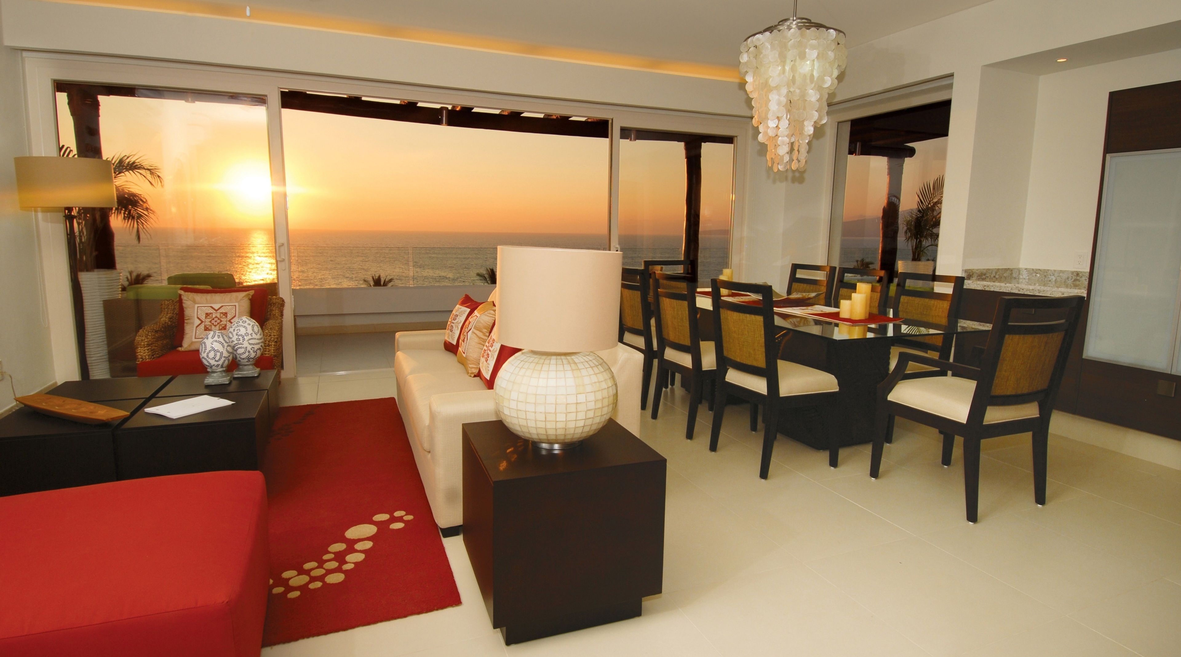 A modern living room with a large window overlooking the sea, a dining area with a chandelier, and a cozy seating area with a red sofa and a decorative lamp.