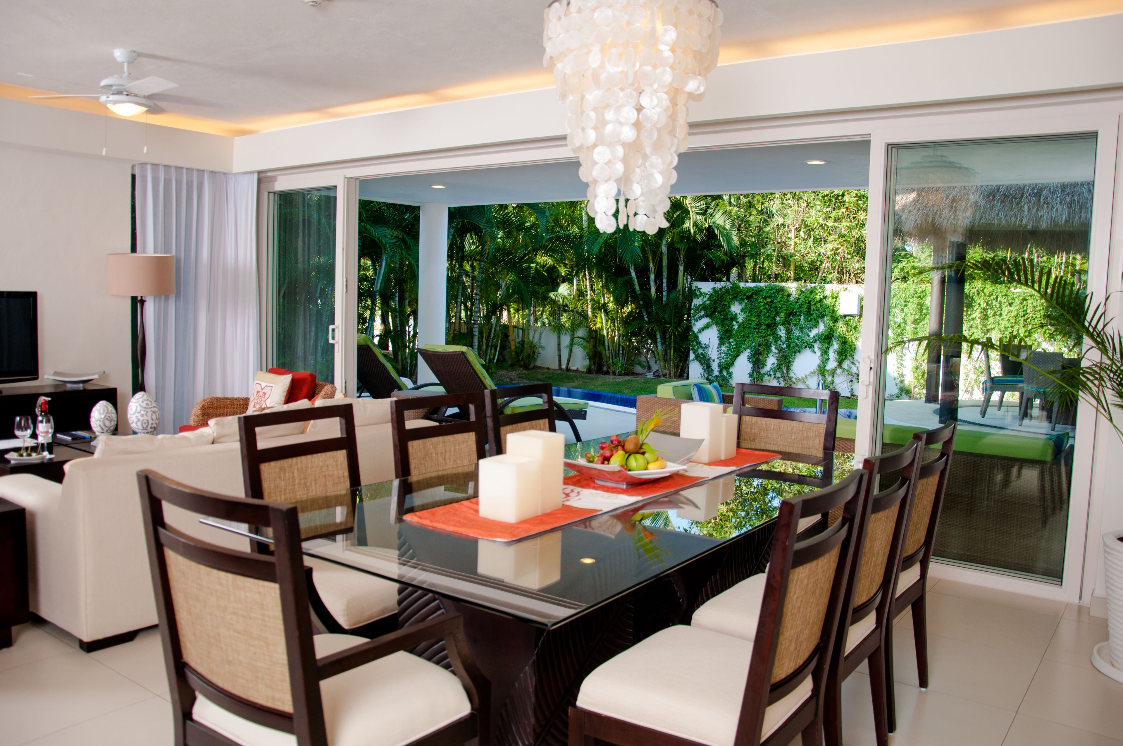 A modern dining area with a glass table, chairs, and a view of a swimming pool and greenery.
