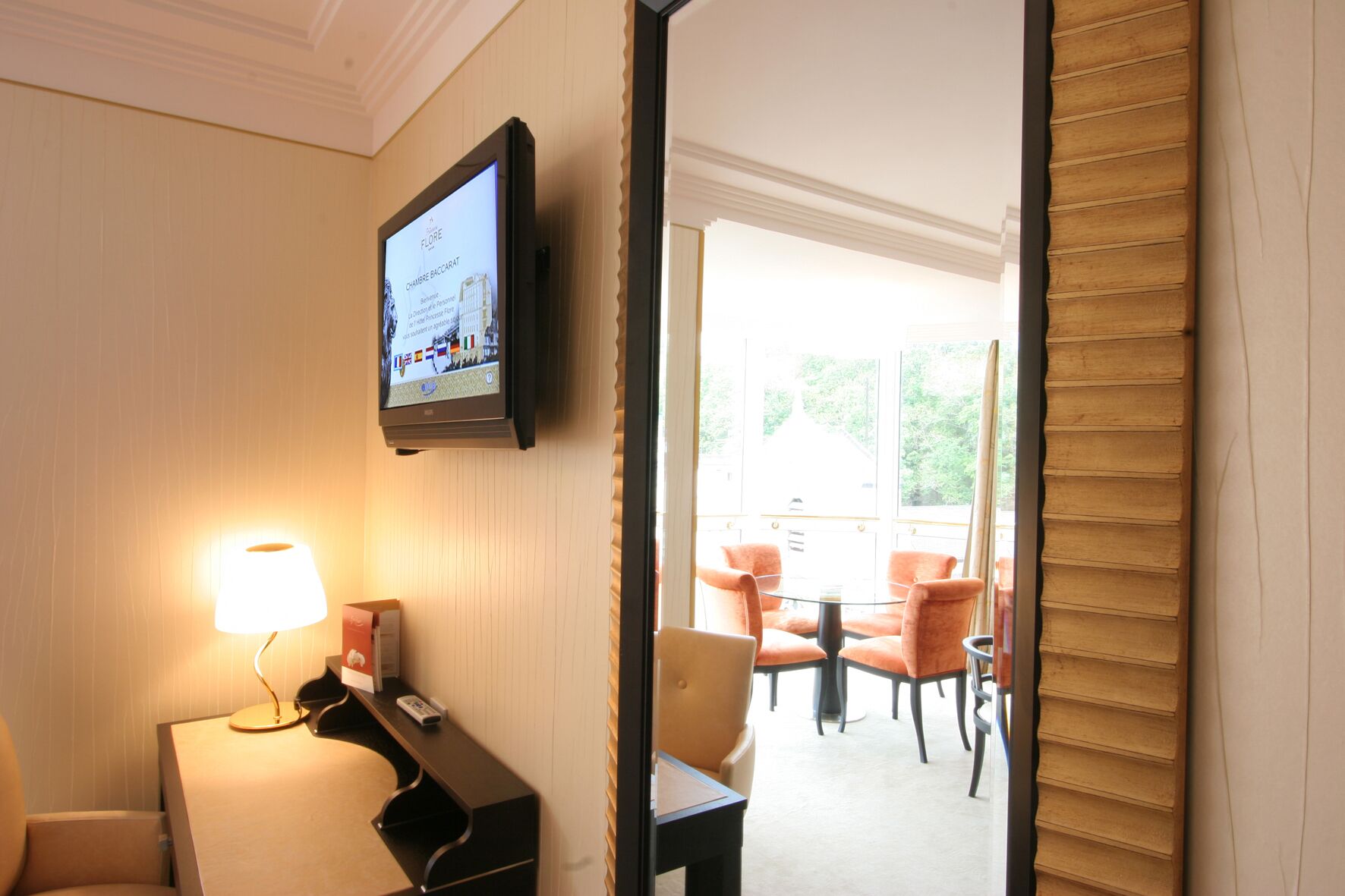 Habitación de hotel con un televisor montado en la pared, un escritorio con lámpara y vista al área de estar exterior a través de una puerta abierta.