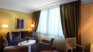A hotel room with a sofa, armchair, and a small table with a vase. There is a window with curtains and a painting on the wall.