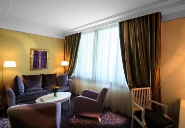 A hotel room with a sofa, armchair, and a small table with a vase. There is a window with curtains and a painting on the wall.