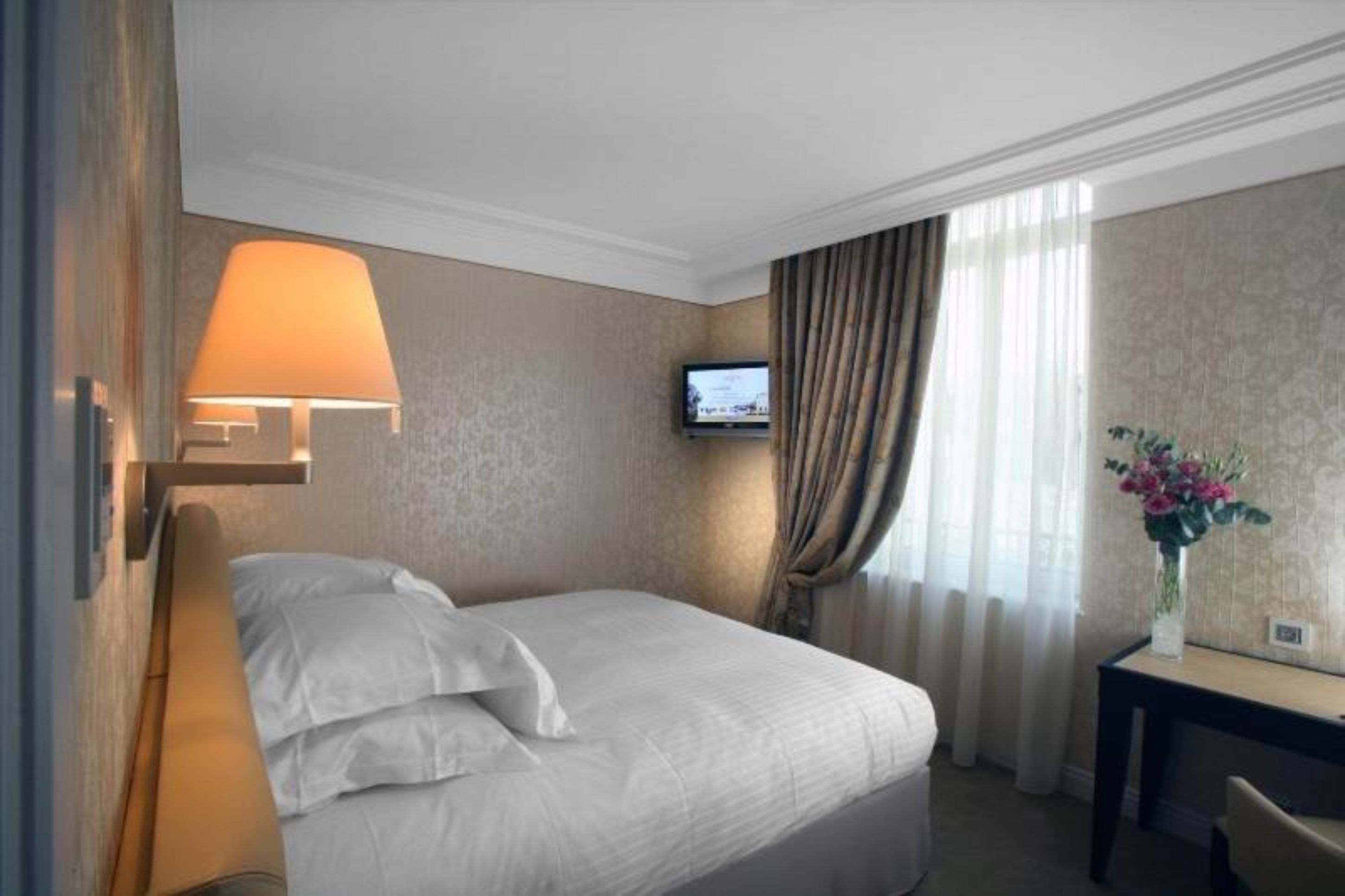 Habitación de hotel con cama, escritorio, lámpara, televisor y un florero con flores.