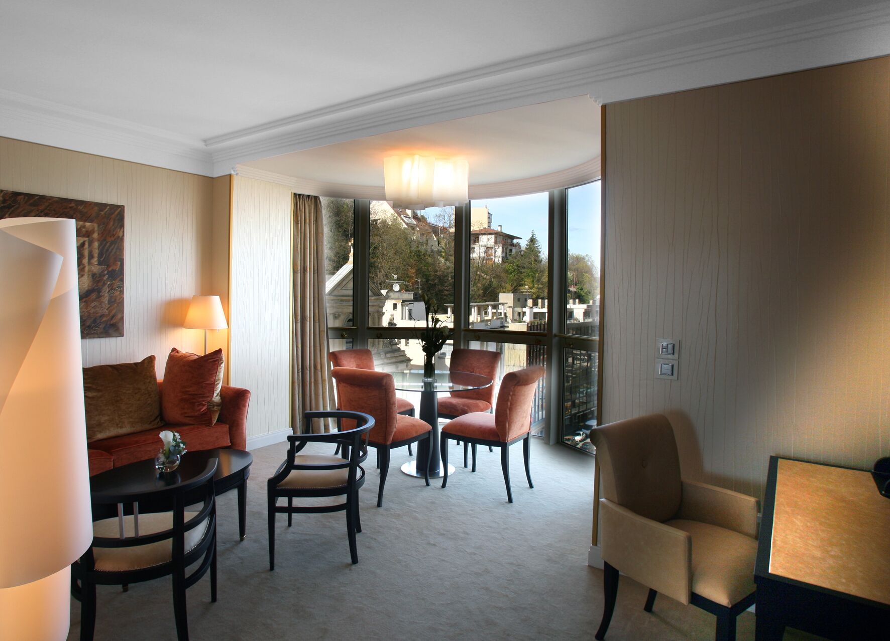 A hotel room with a sofa, two armchairs, a small table, and a view of buildings through large windows.