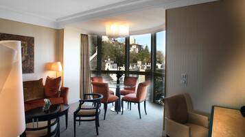 A hotel room with a sofa, two armchairs, a small table, and a view of buildings through large windows.