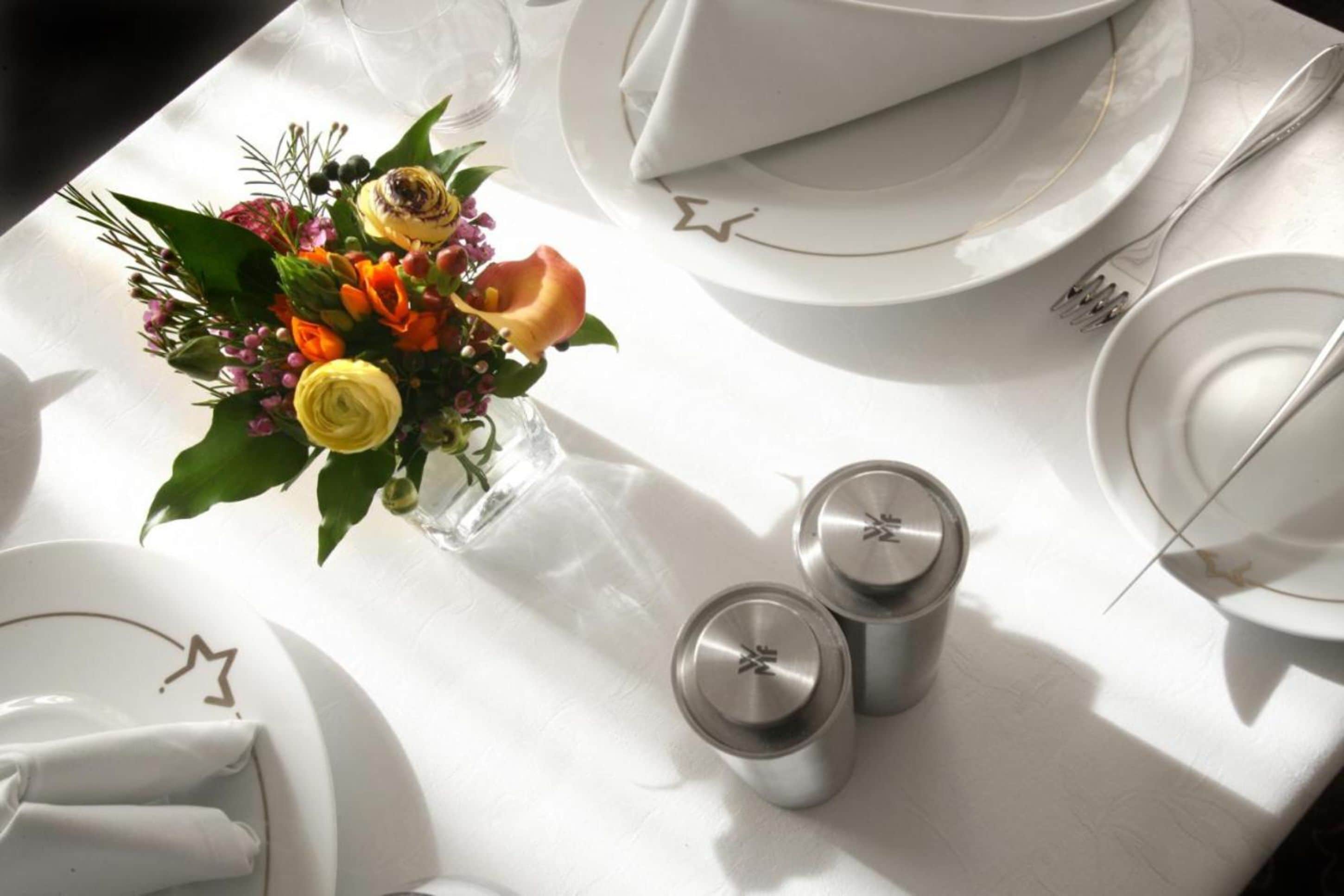 Une table dressée avec de l’argenterie blanche, des serviettes pliées et un vase en verre rempli de fleurs.