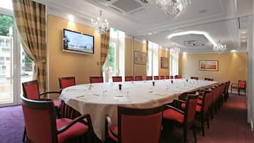 A conference room with a long table, red chairs, chandeliers, and a television screen displaying a welcome message.