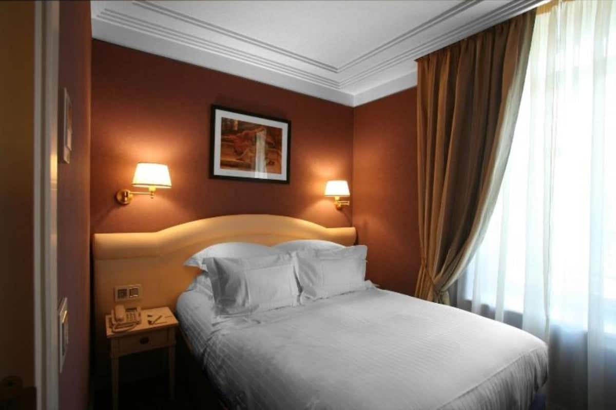 A hotel room with a bed, two bedside lamps, a nightstand with a phone, and a painting on the wall.