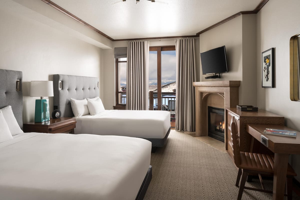 A hotel room with two beds, a desk, a TV, a fireplace, and a balcony with a view of the mountains.