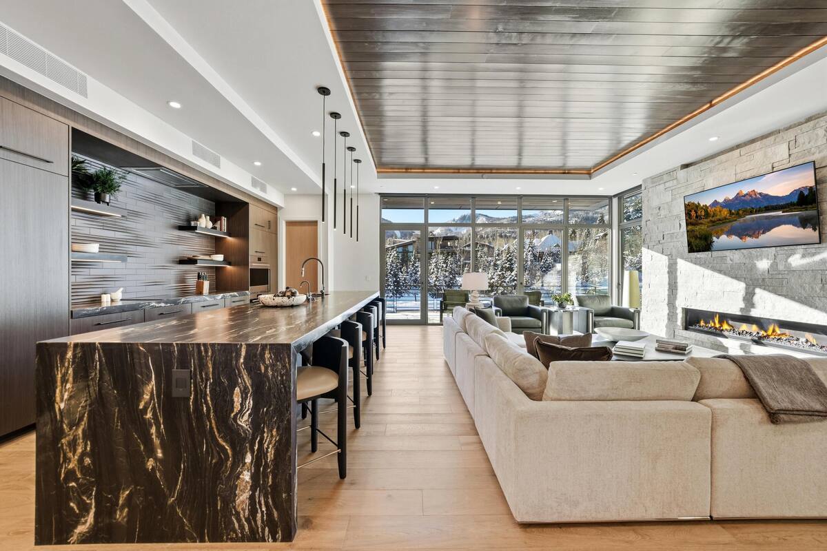 A modern kitchen with a marble island, a dining area with bar stools, a large sofa, and a fireplace.