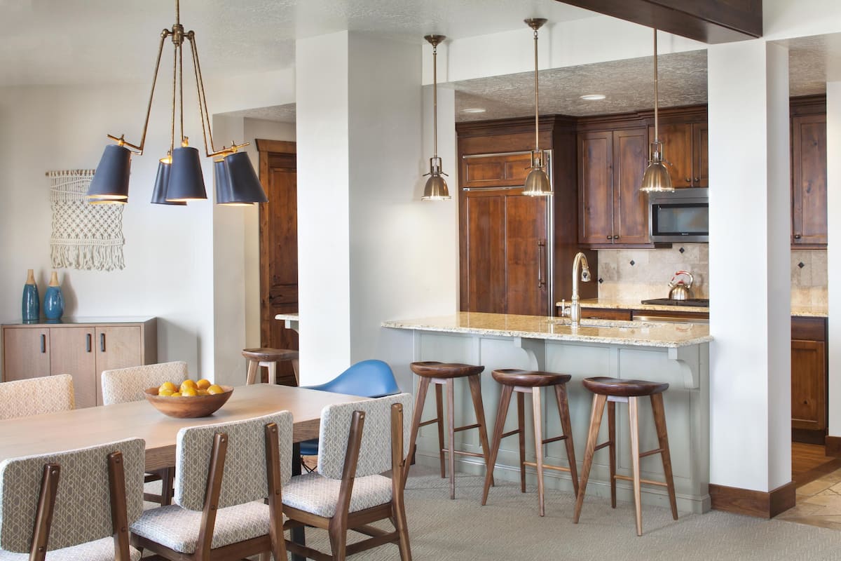 A modern kitchen with wooden cabinets, a central island with stools, pendant lighting, and a dining area with a table and chairs.