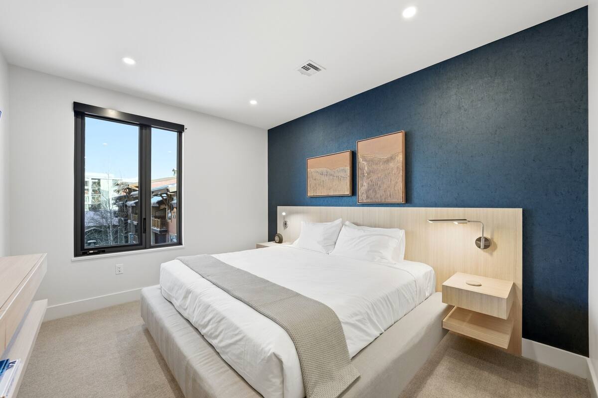 A modern bedroom with a large bed, a bedside table, a nightstand, a window, and two wall-mounted artworks.