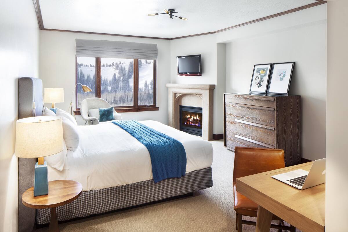 A hotel room with a bed, a desk with a laptop, a fireplace, a TV, and a window with a view of trees.