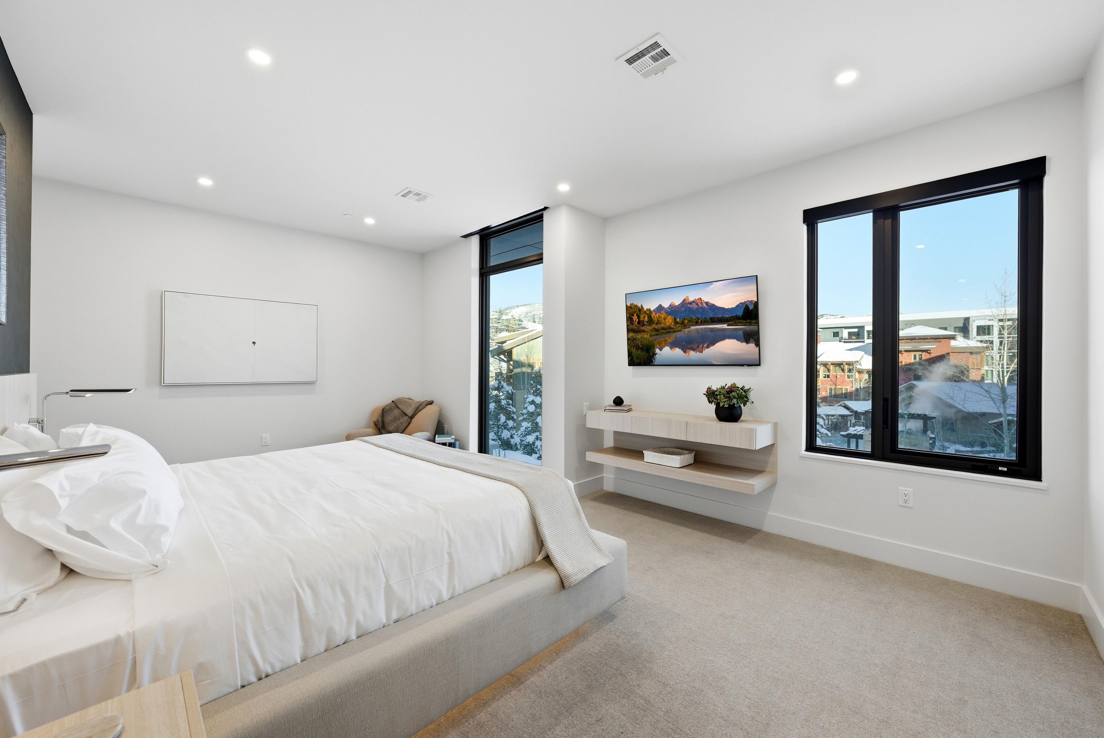Un dormitorio moderno con una cama grande, una mesita de noche, un estante pequeño con una planta y una ventana con vistas a la ciudad.