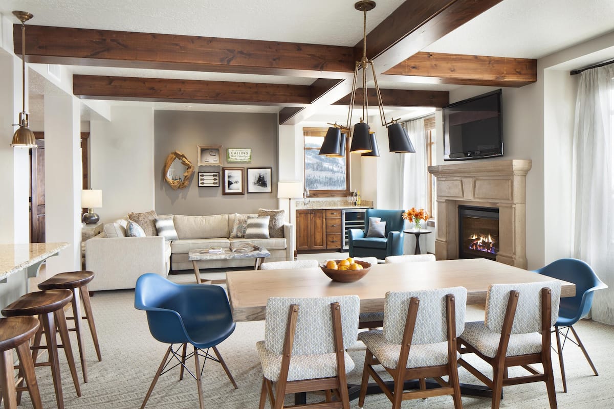 A spacious living room with a large wooden dining table, blue chairs, a fireplace, and a television mounted above it.
